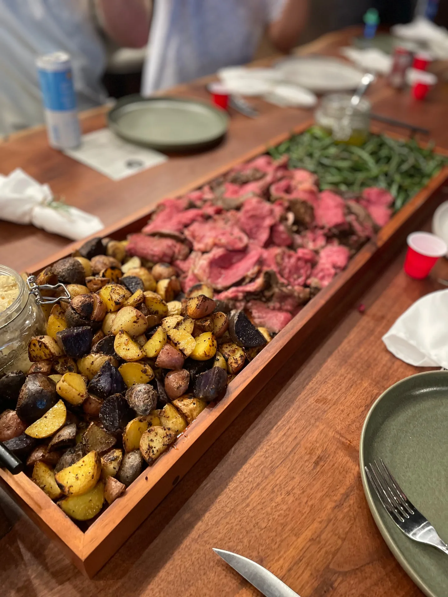 Long wooden serving board of sliced roast beef, roasted potatoes and green beans down the middle of a dinner table